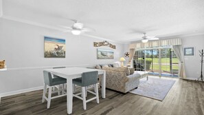 A modern living room with a dining area, featuring a ceiling fan, a beach-themed painting, and a welcome sign. - 613 Wedgewood - Barefoot Resort Rentals (North Myrtle Beach)