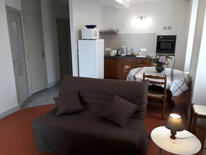 Een knus woongedeelte met een bank, een kleine tafel met lamp, een kitchenette en een eethoek.