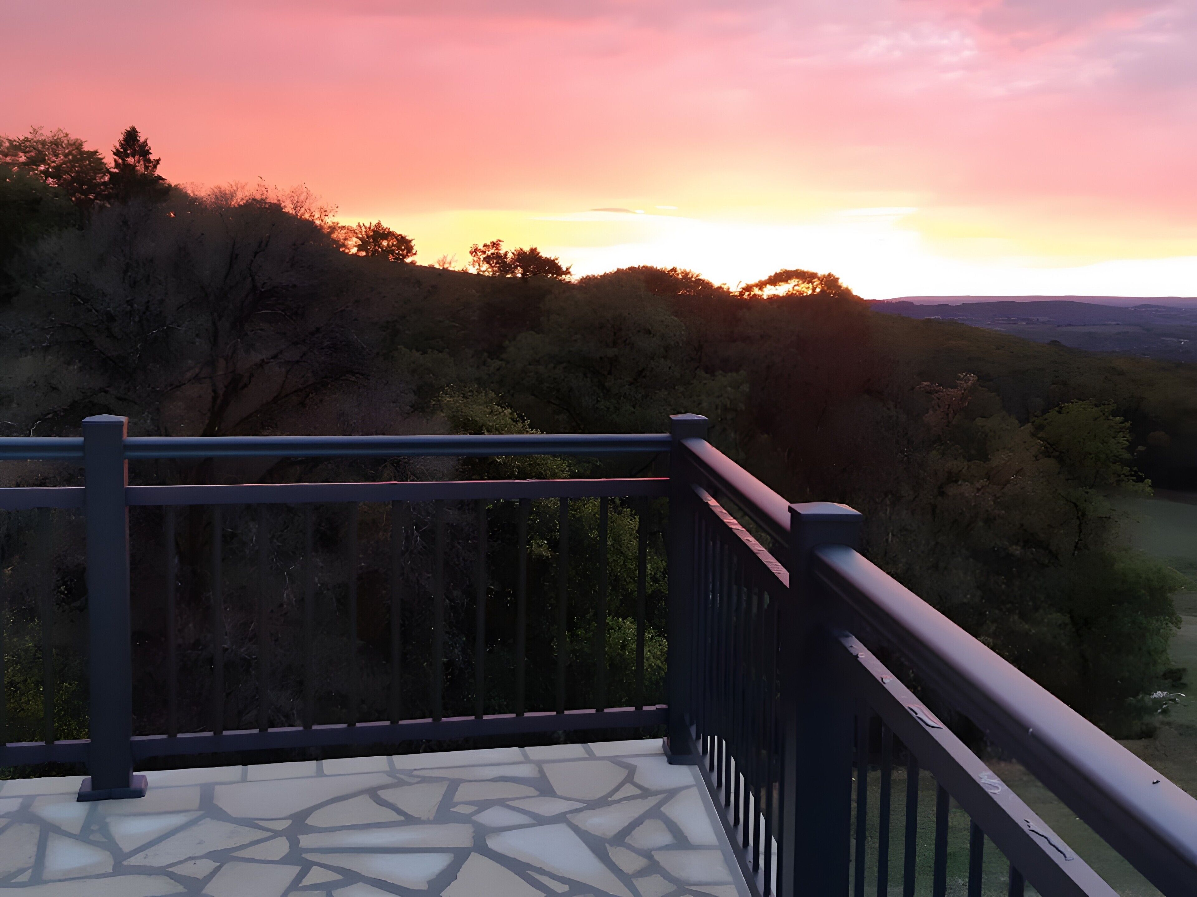 A balcony with a stone floor and metal railing overlooking a landscape with trees and a sunset.