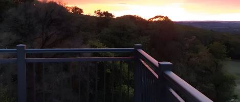 A balcony with a stone floor and metal railing overlooking a landscape with trees and a sunset.