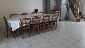 Un comedor con una mesa preparada para seis personas, rodeada de sillas de madera, un jarrón con flores y una zona de cocina con un refrigerador y una escalera de madera.