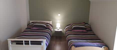 A small, single-room accommodation with two twin beds, a bedside table with a lamp, and a window providing natural light.