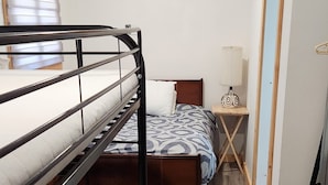 Une chambre avec des lits superposés, une table de chevet avec une lampe, un plancher en bois et une fenêtre avec des stores.