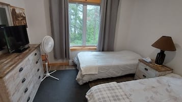 A bedroom with a TV, a fan, a lamp, two beds, a dresser, and a window with a view of trees.