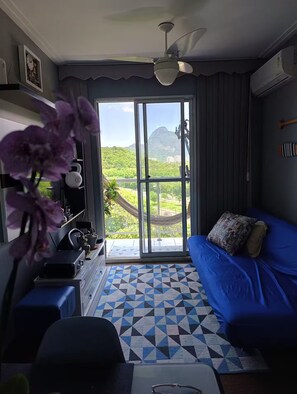 A room with a blue sofa, a patterned rug, a ceiling fan, and a view of greenery and mountains through a large window. - Fully equipped apartment (Riocentro / Olympic Park) (Rio de Janeiro)