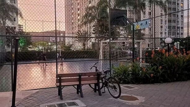 A fenced outdoor area with a bench and a bicycle, surrounded by greenery and buildings in the background. - Fully equipped apartment (Riocentro / Olympic Park) (Rio de Janeiro)