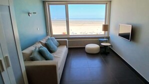 A room with a sofa, a small round table, a radiator, a TV, and a window with a beach view. - Beachfront Knokke-Heist Studio w/ Oceanfront Views (Knokke-Heist)