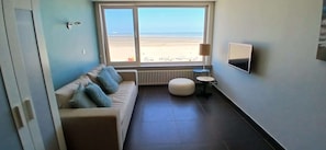 A room with a sofa, a small round table, a radiator, a TV, and a window with a beach view. - Beachfront Knokke-Heist Studio w/ Oceanfront Views (Knokke-Heist)