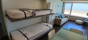 A bunk bed with a mattress on the top bunk and a single bed below. There is a sofa with cushions and a view of the beach through a window. - Beachfront Knokke-Heist Studio w/ Oceanfront Views (Knokke-Heist)