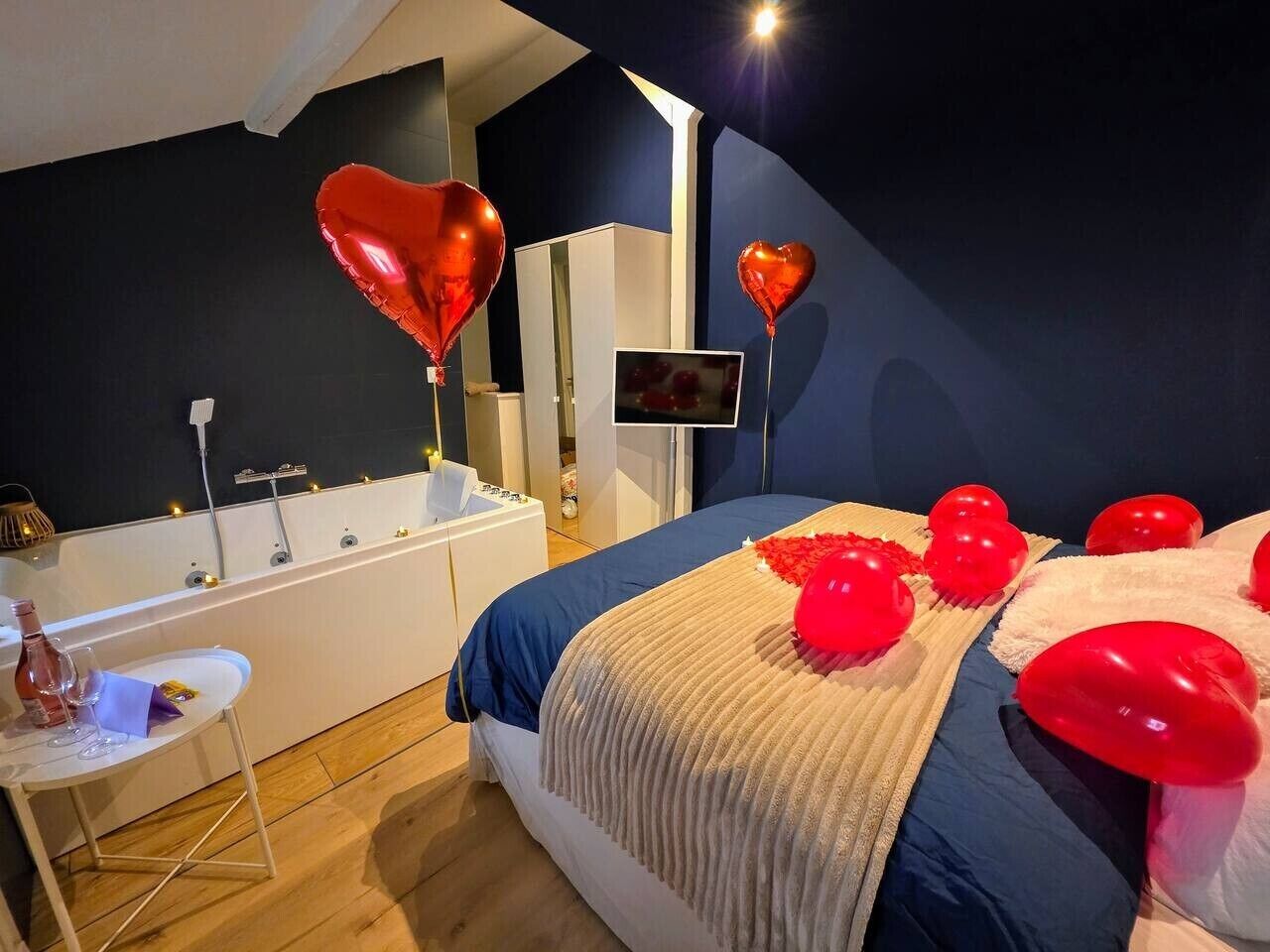 Ein Schlafzimmer mit einem Bett, einer Badewanne, einem kleinen Tisch mit einer Flasche und Gläsern sowie herzförmigen Ballons.