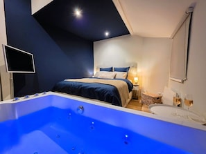 A modern bedroom with a large bathtub, a bed with dark bedding, a bedside table with a lamp, and a wall-mounted TV. - Spa Suite 15 minutes from Strasbourg (Schiltigheim)