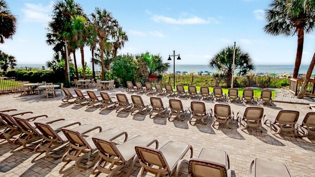 Ein sonniger Außenbereich mit Liegestühlen und Tischen, der einen Blick auf einen Strand und Palmen bietet.