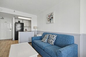 A living room with a blue sofa, a white coffee table, and a kitchen in the background. - Oceanfront 1BR Suite + Beach Access + Lazy River (Myrtle Beach)
