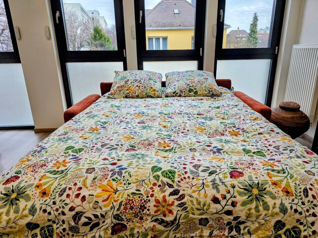 Ein Bett mit floralem Überwurf, ein Fenster mit Blick auf Gebäude und eine Heizung.