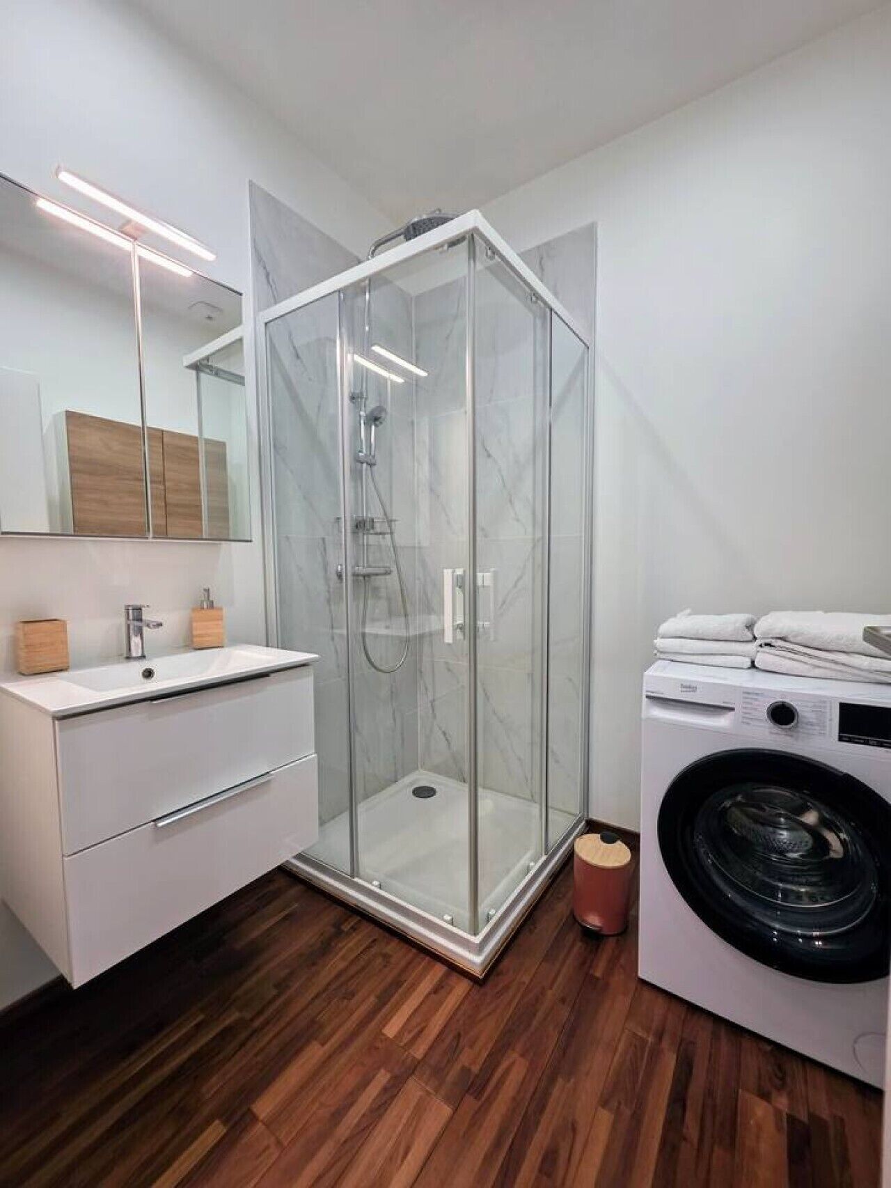 Ein modernes Badezimmer mit einer gläsernen Duschkabine, einem Waschbecken mit Holzschrank und einer Waschmaschine mit Frontlader.