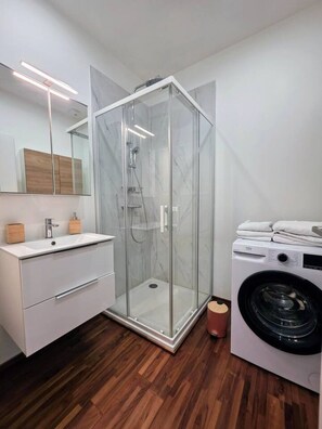 Ein modernes Badezimmer mit einer gläsernen Duschkabine, einem Waschbecken mit Holzschrank und einer Waschmaschine mit Frontlader.