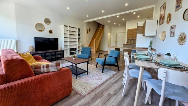 Een moderne woonkamer met een bank, fauteuils, een salontafel, een televisie en een trap die naar een keuken leidt.