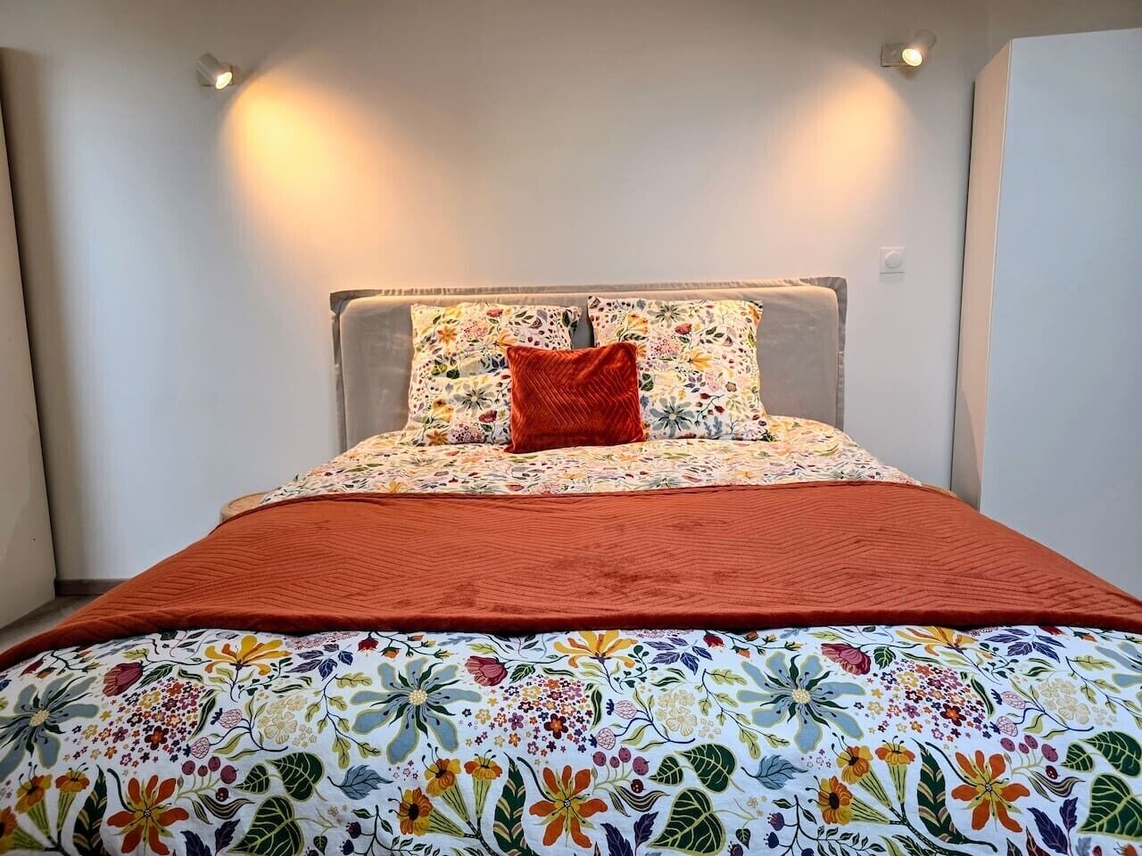 Ein Bett mit blumigem Überwurf und roter Decke, ein Kissen mit Blumenmuster sowie ein Kopfteil mit Leuchte.