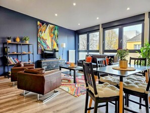 A modern living room with a sofa, dining table, and chairs, a bookshelf, and a large abstract painting. - Charming 55-square-meter duplex, 15 minutes from Strasbourg (Schiltigheim)