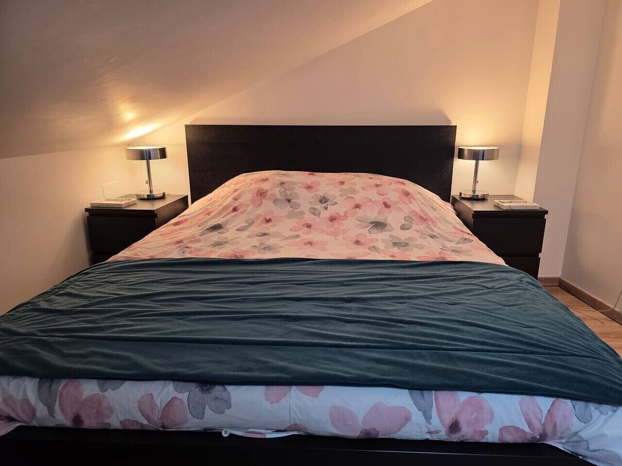 Una cama con una colcha floral, dos lámparas de noche y un cabecero oscuro en una habitación con paredes lisas.