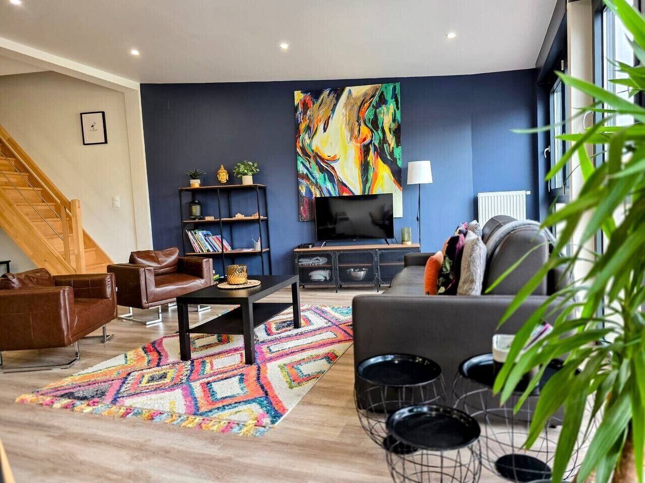 A modern living room with a staircase, a large abstract painting, a flat-screen TV, and a variety of seating options.