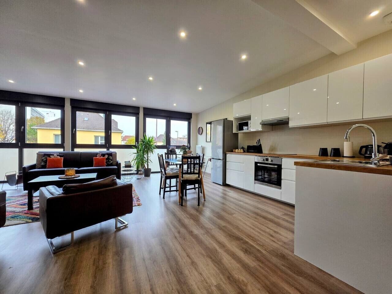 Eine moderne Küche mit weißen Schränken, ein Essbereich mit einer schwarzen Couch und Holzstühlen, sowie ein großes Fenster für natürliches Licht.