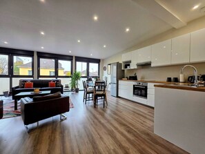 Una cocina moderna con armarios blancos, un comedor con sofá negro y sillas de madera, y un amplio ventanal que proporciona luz natural.