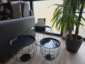 Una sala de estar moderna con un sofá gris, tres mesas de centro de estructura metálica, una planta en maceta y un ventanal con vistas.
