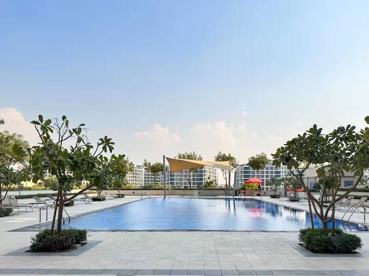 Una piscina con tumbonas, una zona con sombra y un edificio al fondo.