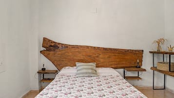Un dormitorio con cabecera de madera, edredón estampado con motivos florales, lámpara de noche y una pequeña estantería con adornos.