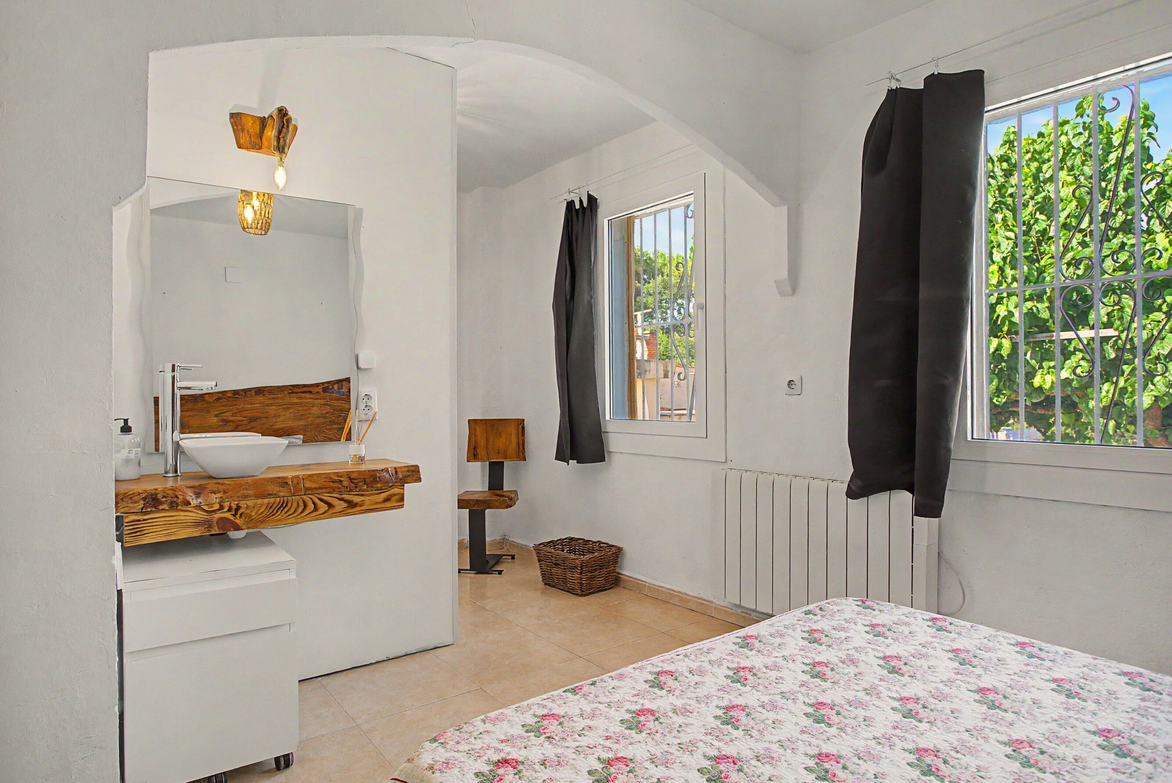 A bedroom with a wooden vanity, a bed with a floral bedspread, a window with a view of greenery, and a radiator.