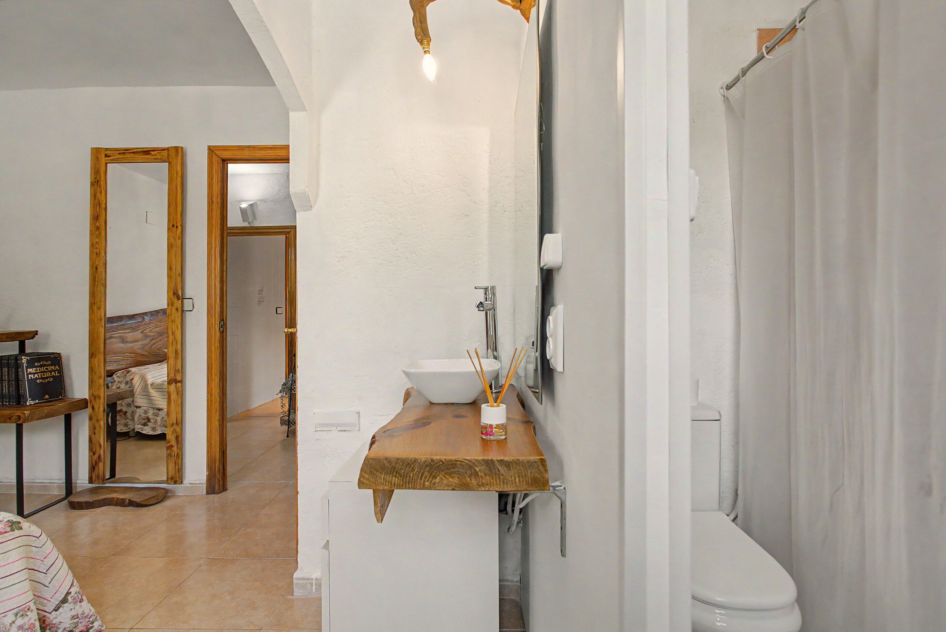 Een badkamer met een houten wastafelmeubel, een wastafel en een toilet. Boven de wastafel hangt een spiegel en rechts is er een douchegordijn.