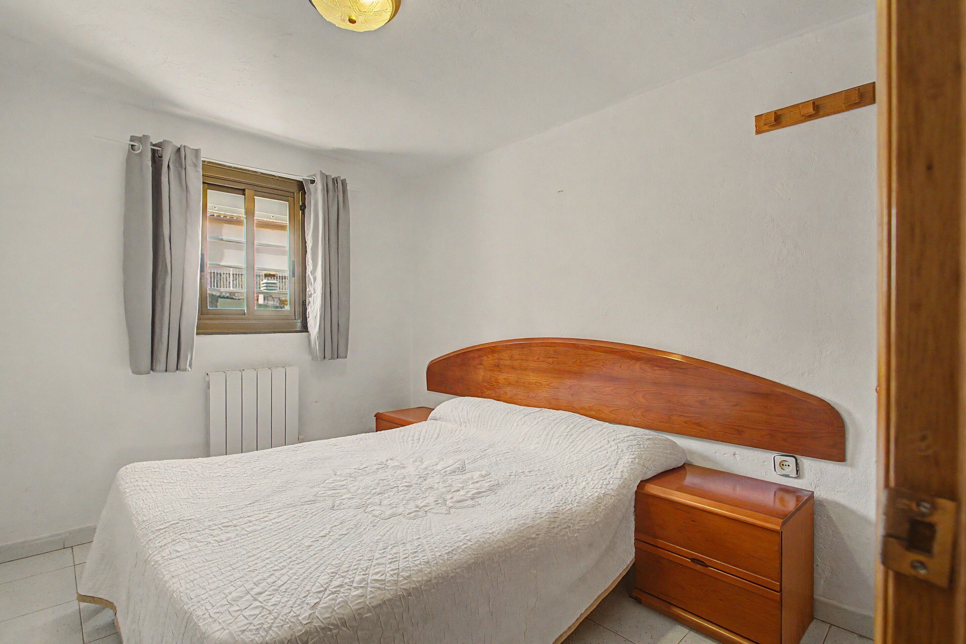 Un dormitorio con cama individual, cabecera de madera, mesita de noche, ventana con cortinas, radiador y puerta de madera.