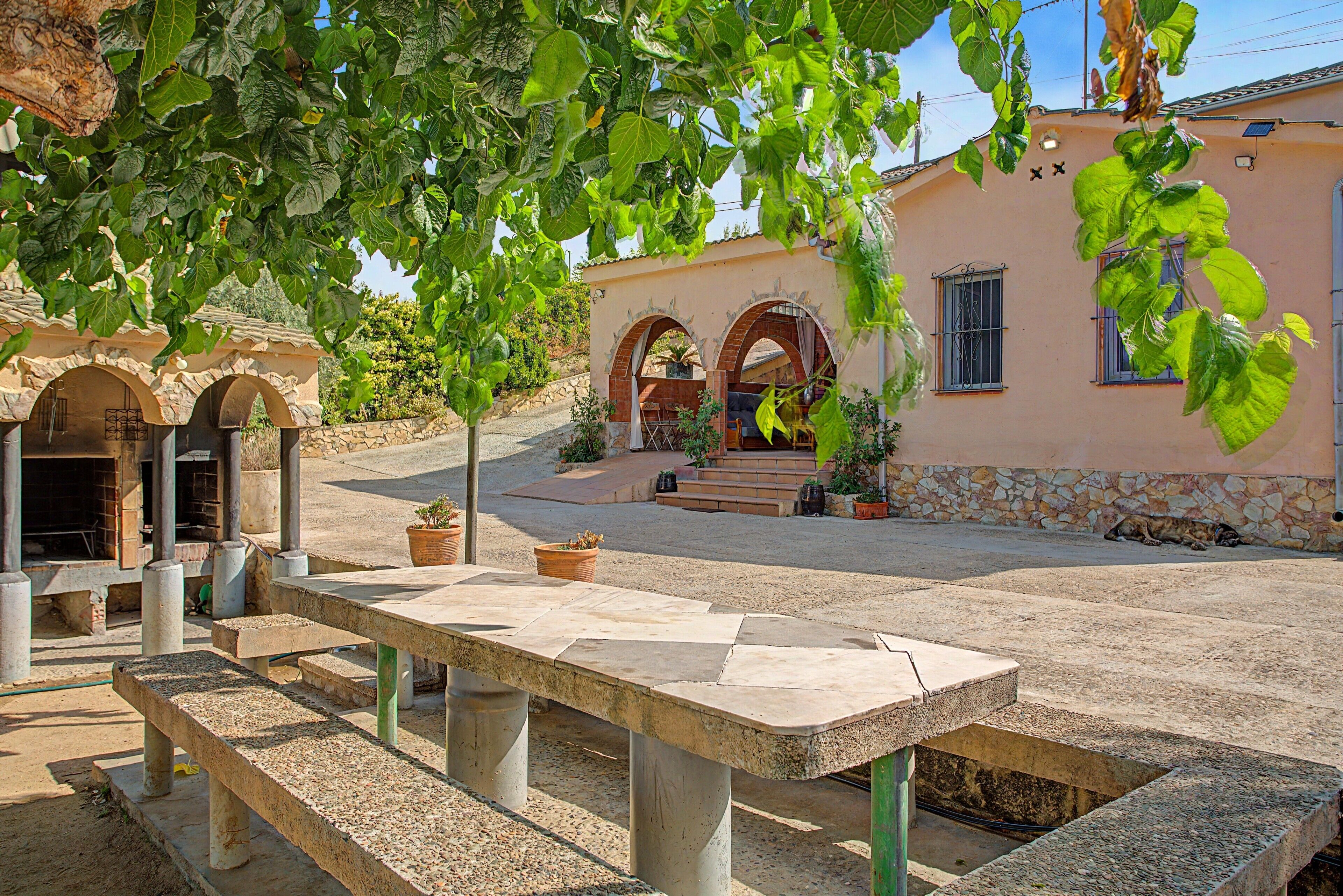 Un patio al aire libre con una mesa de piedra y bancos, rodeado de abundante vegetación y un edificio con arcos y techo de tejas.