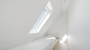 Een kleine, witte kamer met een enkel bed, een schuine kap en een dakraam.
