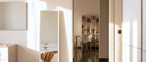 A modern hotel room with a bed, a desk, a chair, and a bookshelf.
