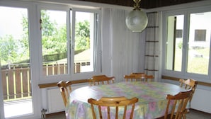 A dining room with a round table and six wooden chairs, large windows, and a hanging light fixture.