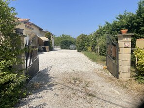 Une allée de gravier mène à une entrée fermée par un portail, avec une maison située sur la gauche.