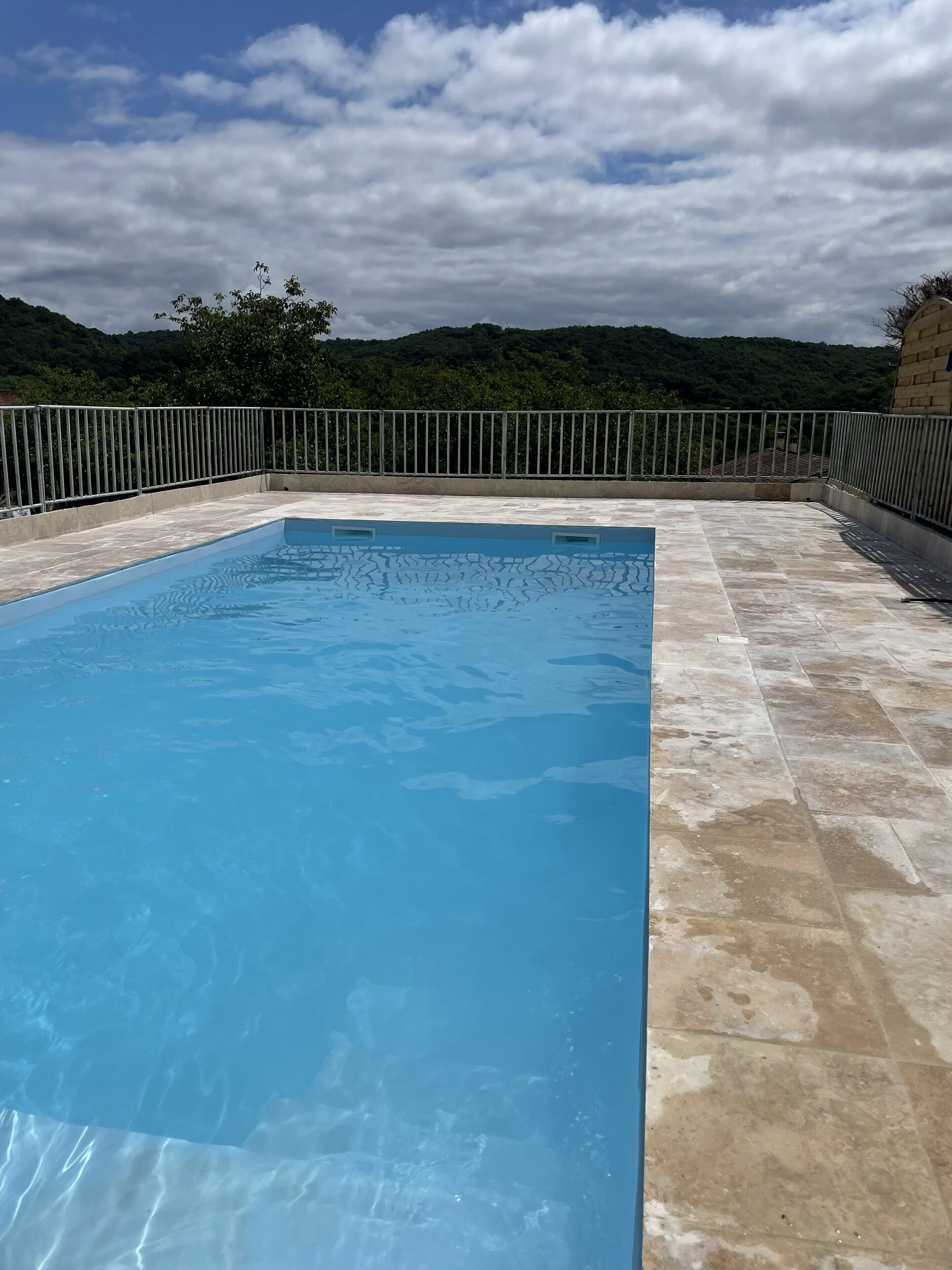 A rectangular swimming pool with a tiled deck, surrounded by a metal fence, set against a backdrop of green hills and a partly cloudy sky.