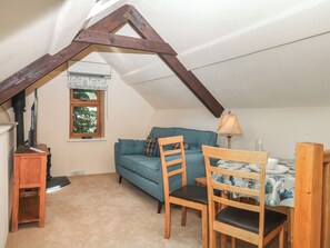 A cozy attic bedroom with a sloped ceiling, a blue sofa, a wooden dining table, and a small wooden heater. - Lustleigh Mill Barn (Newton Abbot)