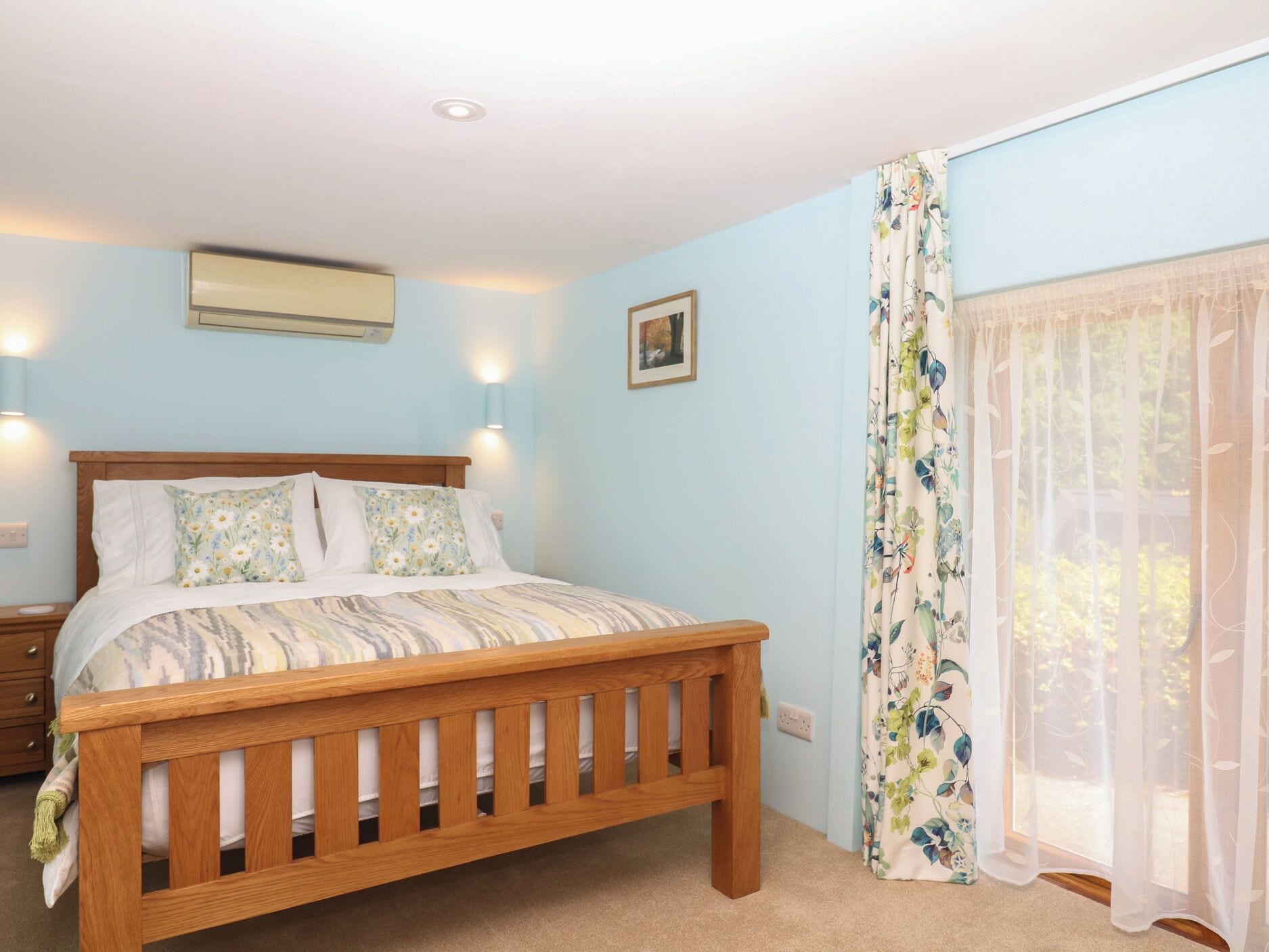 Un dormitorio con cama de madera, una mesita de noche, aire acondicionado montado en la pared y una ventana con cortinas florales.