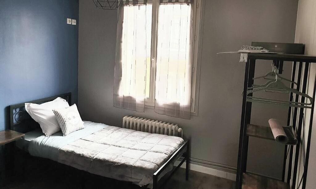 A bedroom with a bed, a radiator, a window with curtains, a small table, and a wall-mounted shelf with a towel and a hanger.