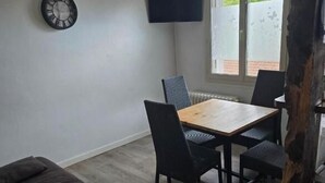 Een kamer met een houten eettafel, zwarte stoelen, een donkere bank, een wandklok en een aan de muur bevestigde televisie.