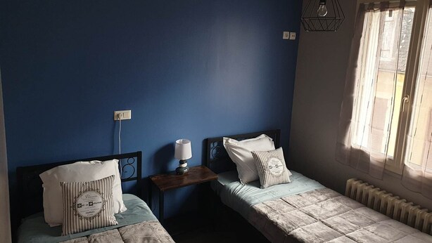 Een kamer met twee bedden, een nachtkastje met een lamp, een radiator en een raam met gordijnen.