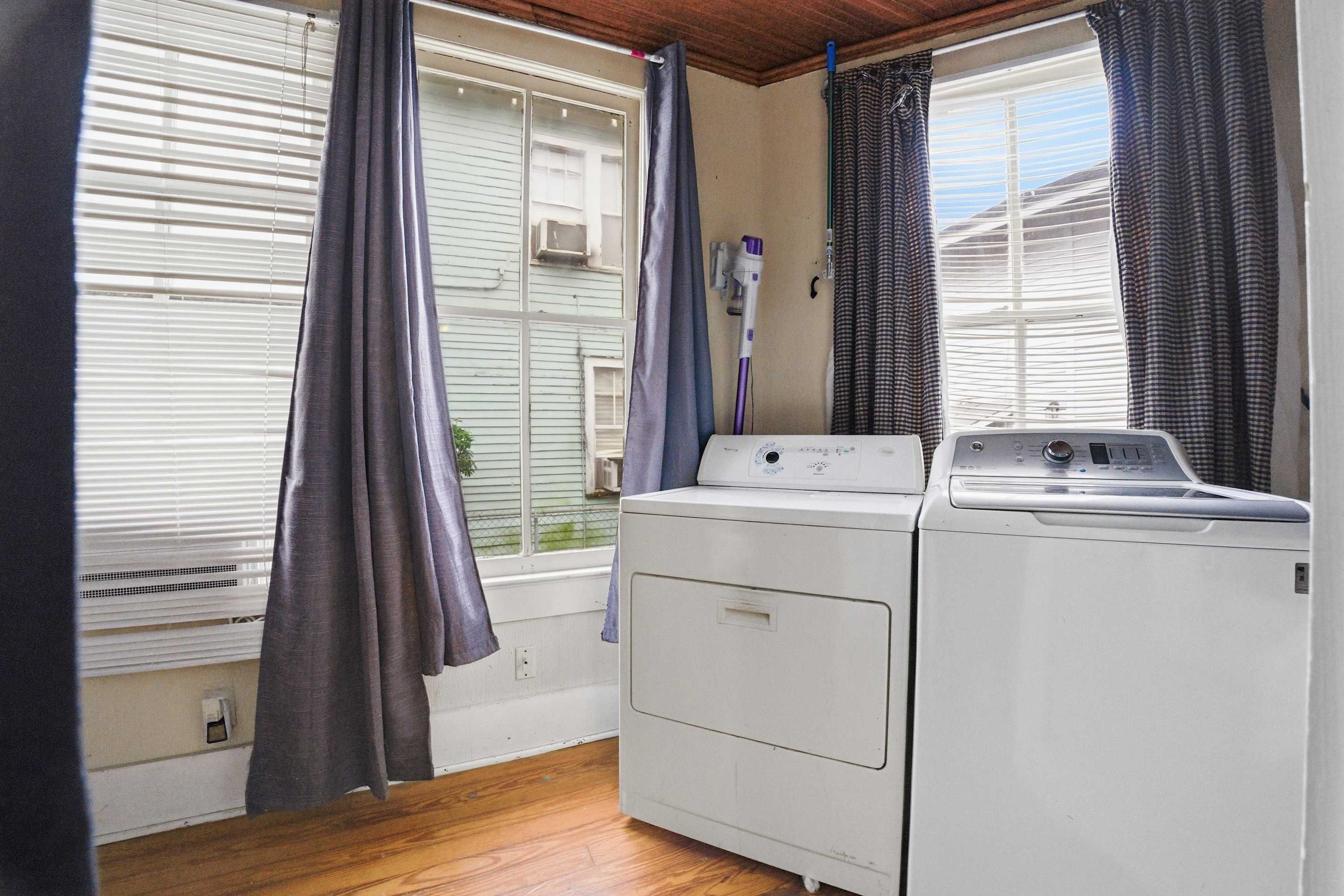 Une buanderie équipée d’un lave-linge et d’un sèche-linge, avec un plancher en bois et une vue sur une maison par une fenêtre dotée de stores.