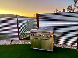Una parrilla exterior con panel de control, frente a una pared de metal corrugado y una vista de las montañas al fondo.