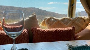 Et værelse med udsigt til bjerge gennem et vindue, et glas vin på et bord og et hyggeligt siddeområde med puder.