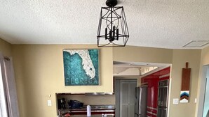 A dining area with a glass table, blue chairs, a kitchen in the background, and a ceiling light fixture. - Siesta Beach Getaway With Heated Pool (Siesta Key)