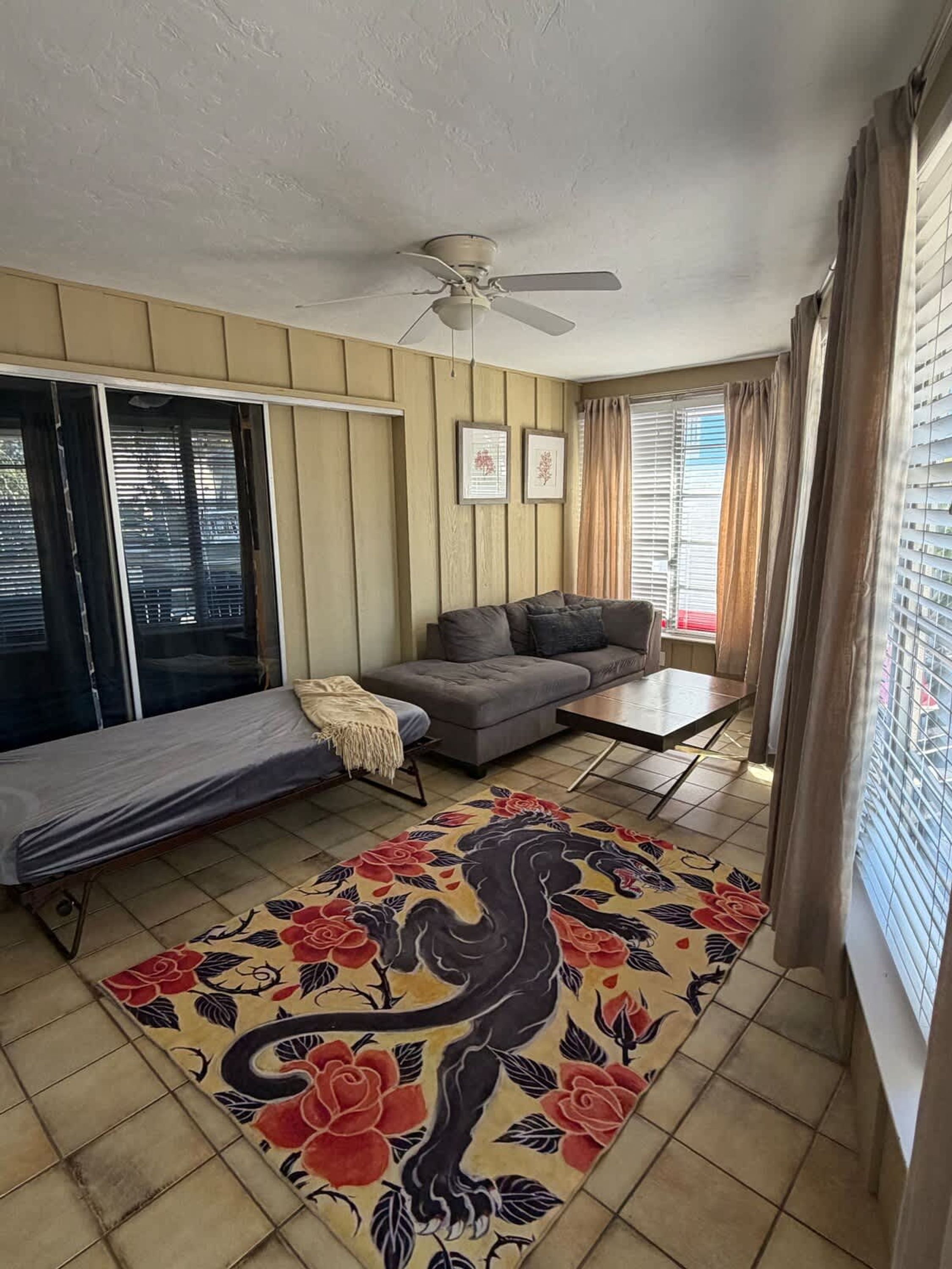 A room with a sofa, a small table, a ceiling fan, and a bed with a colorful rug.
