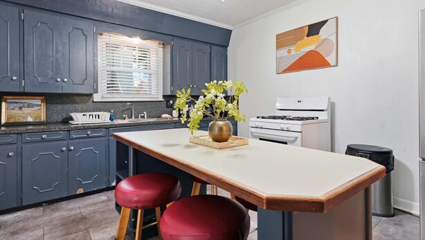 A kitchen with dark blue cabinets, a white countertop, a stove, and a sink. There are two red stools and a vase with flowers on the counter. - Private Bedroom in Spacious Home Near Downtown Laf (Lafayette)